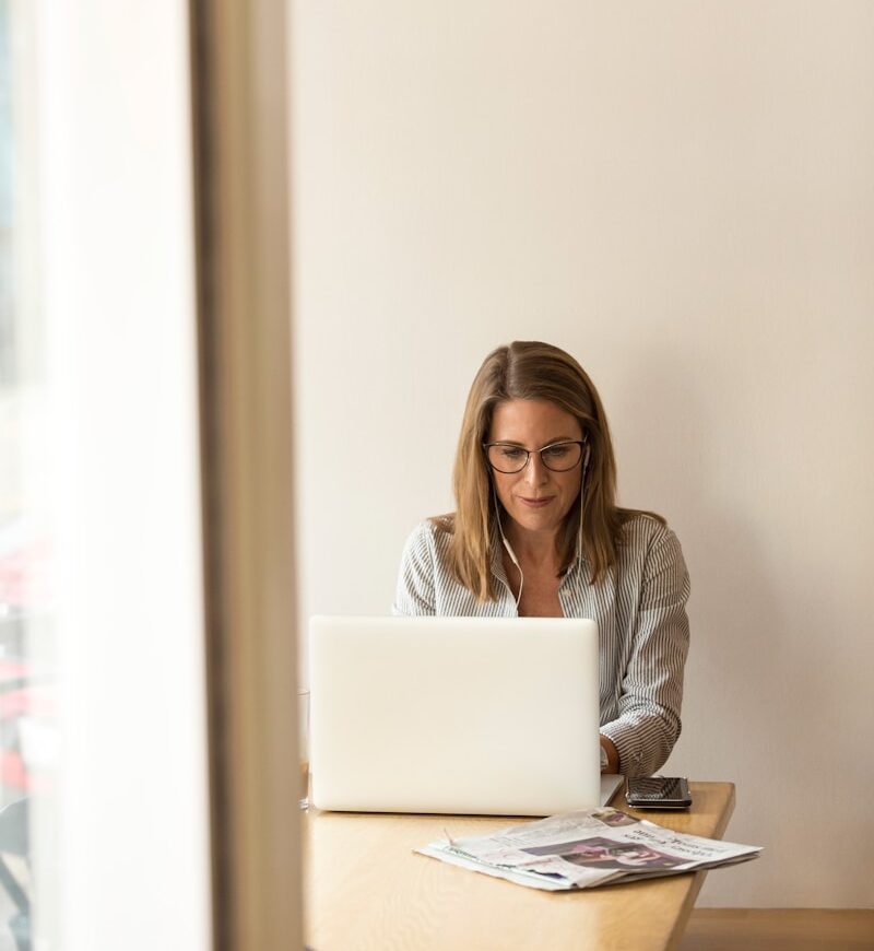 woman wearing grey striped dress shirt sitting down near brown wooden table in front of white laptop computer